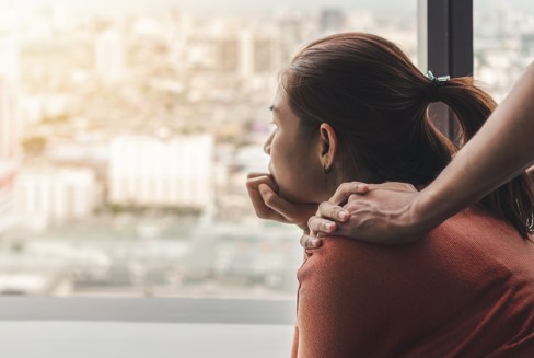 depressed woman staring out window with a hand on her shoulder